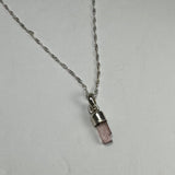 Silver necklace with a small natural pink tourmaline crystal pendant in a cylindrical cap, displayed on a plain light gray surface.