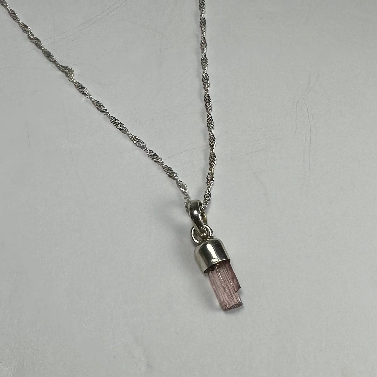 Silver necklace with a small natural pink tourmaline crystal pendant in a cylindrical cap, displayed on a plain light gray surface.