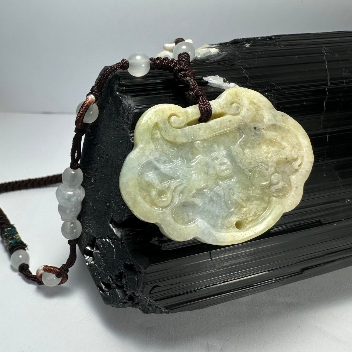 Light green nephrite jade pendant intricately carved with dragon and phoenix motifs, strung on a dark brown cord with translucent beads.