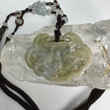 Light green nephrite jade pendant carved with dragon and phoenix motifs, displayed on a clear textured crystal, attached to a dark brown cord with white beads.