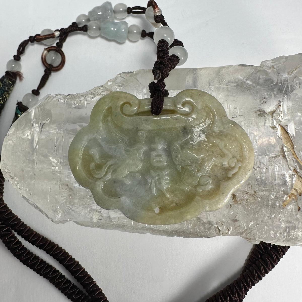 Light green nephrite jade pendant carved with dragon and phoenix motifs, displayed on a clear textured crystal, attached to a dark brown cord with white beads.