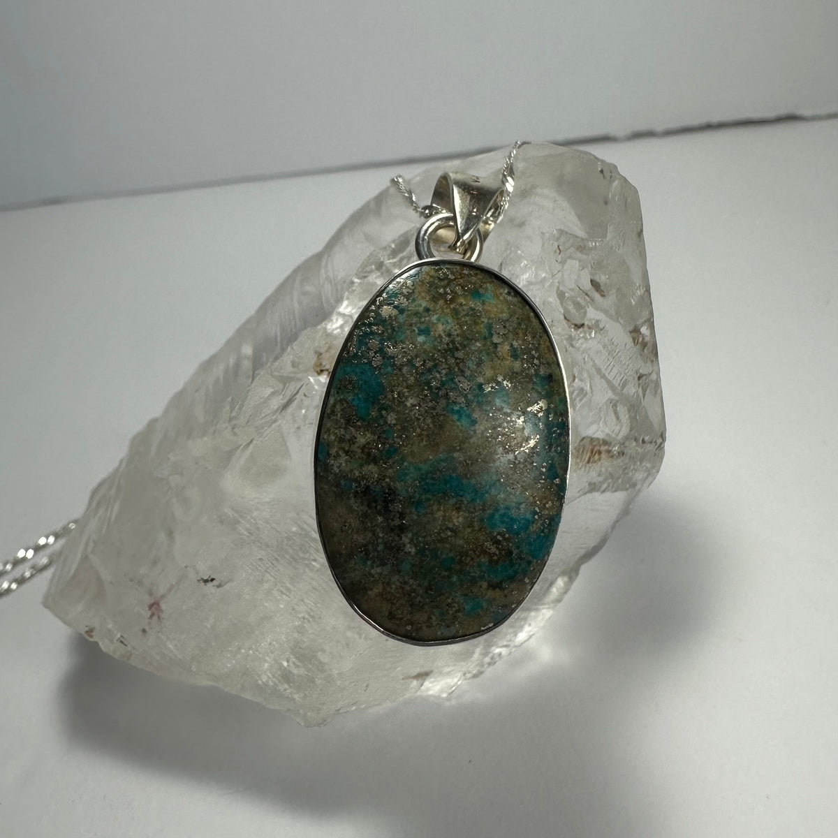Oval turquoise pendant with mottled green and blue stone set in silver, displayed on a rough clear quartz crystal against a light gray background.
