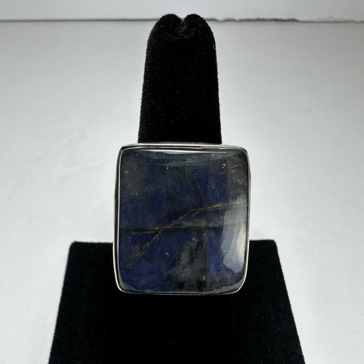 Square labradorite ring with a dark blue and gray iridescent stone set in a simple silver bezel, displayed on a black velvet ring stand.