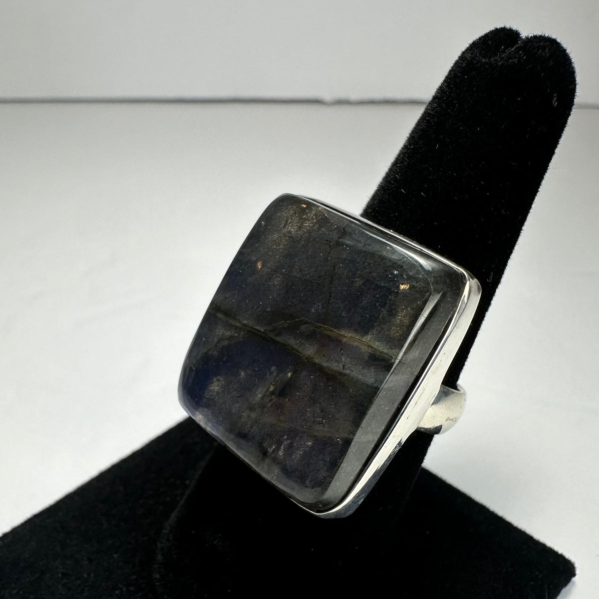 Square labradorite ring with dark blue and gray hues set in a silver band, displayed on a black velvet ring holder against a white background.