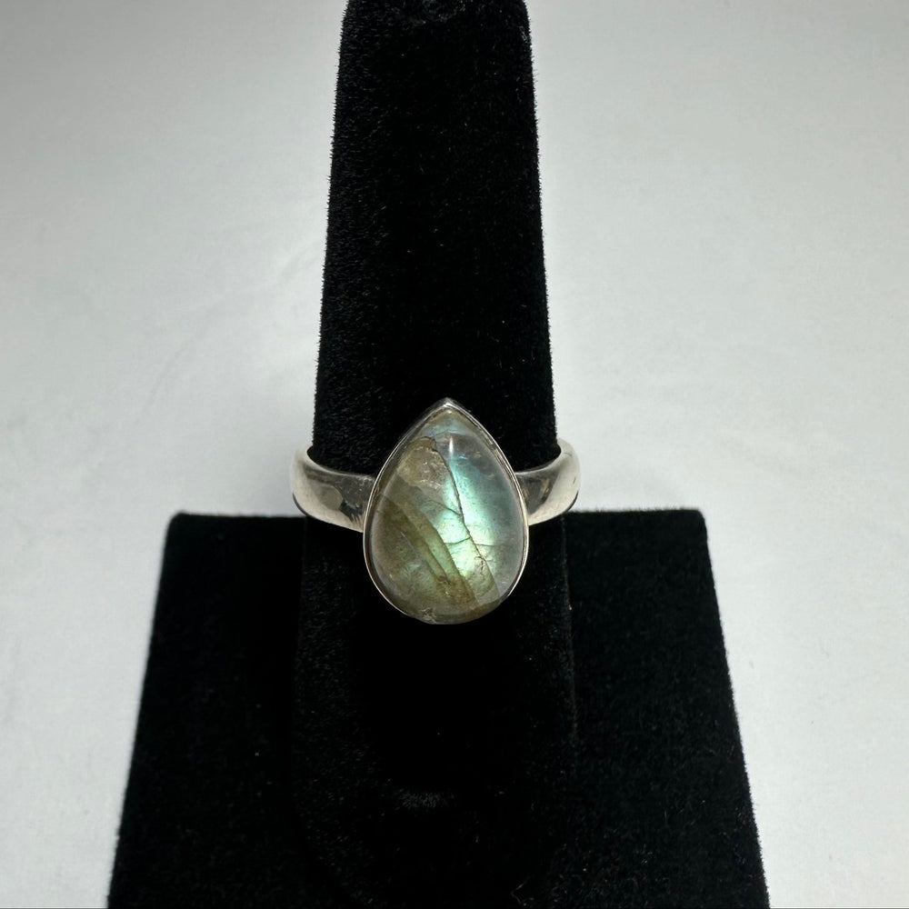 Teardrop-shaped labradorite ring with iridescent blue-green flashes set in a simple silver band on a black velvet ring stand.