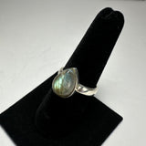 Silver ring with a pear-shaped labradorite stone displaying green and blue iridescence, mounted on a black velvet ring stand against a white background.