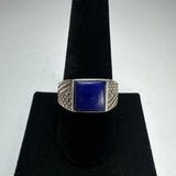 Square lapis lazuli stone set in a silver ring with textured geometric patterns on the band, displayed on a black velvet ring stand.