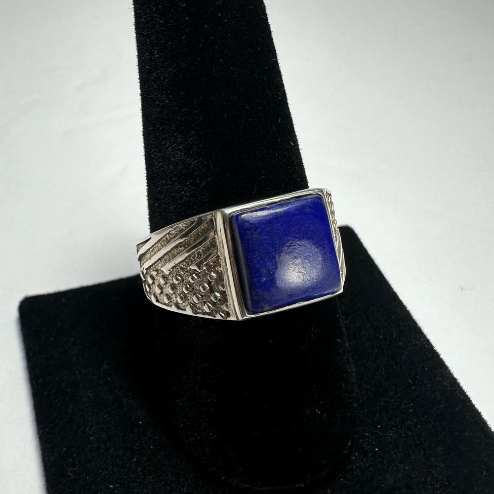 Square lapis lazuli stone set in a silver ring with textured geometric patterns on the band, displayed on a black velvet ring holder.