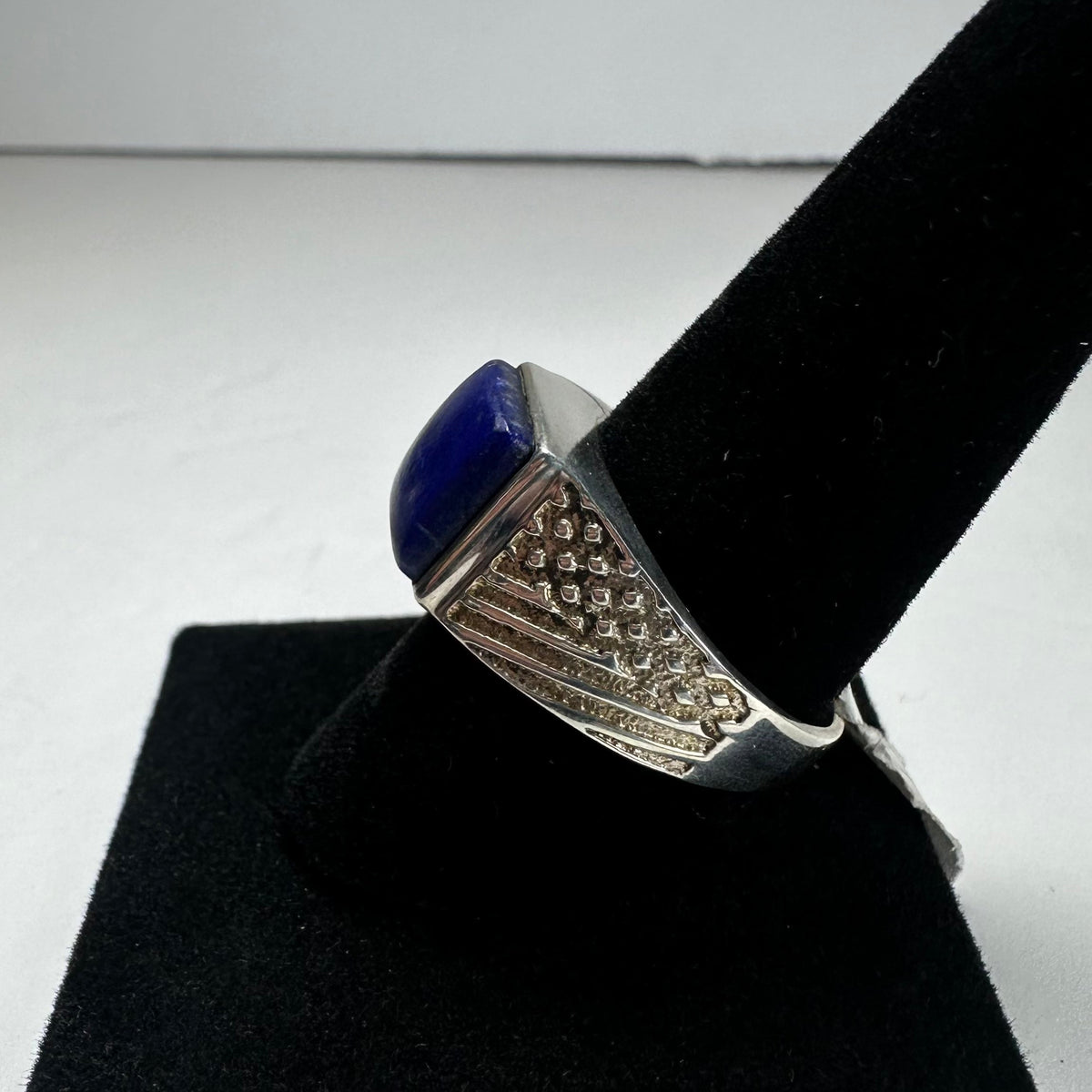 Silver ring with textured geometric pattern on the band, featuring a polished deep blue lapis lazuli stone, displayed on a black velvet ring holder.