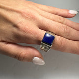 Hand wearing a silver ring with a large, square lapis lazuli stone and intricate textured detailing on the band, shown against a gray background.