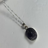 Oval pendant with a rough dark amethyst stone set in a silver bezel on a twisted silver chain, shown on a light gray background.