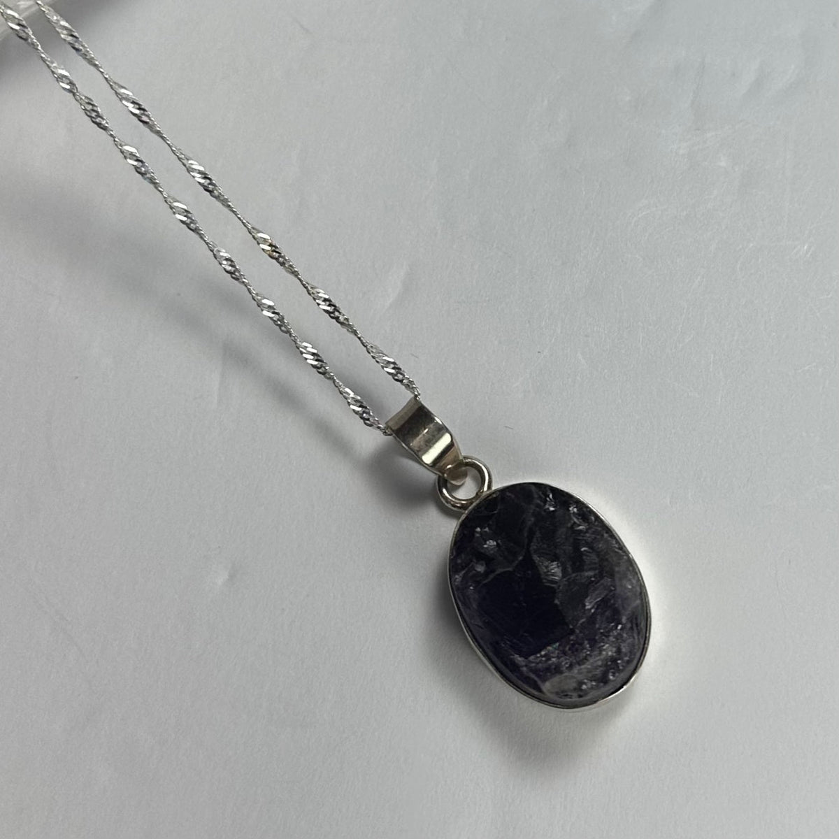 Oval pendant with a rough dark amethyst stone set in a silver bezel on a twisted silver chain, shown on a light gray background.
