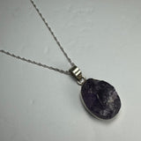 Silver necklace with a natural rough amethyst pendant featuring deep purple and translucent crystal textures, lying on a light gray surface.