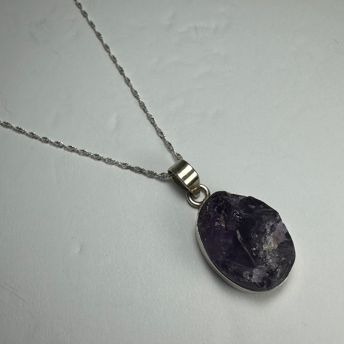 Silver necklace with a natural rough amethyst pendant featuring deep purple and translucent crystal textures, lying on a light gray surface.