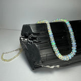 Graduated translucent opal beads with iridescent blue and green flashes strung into a necklace draped over a rough, shiny black tourmaline crystal cluster on a gray surface.