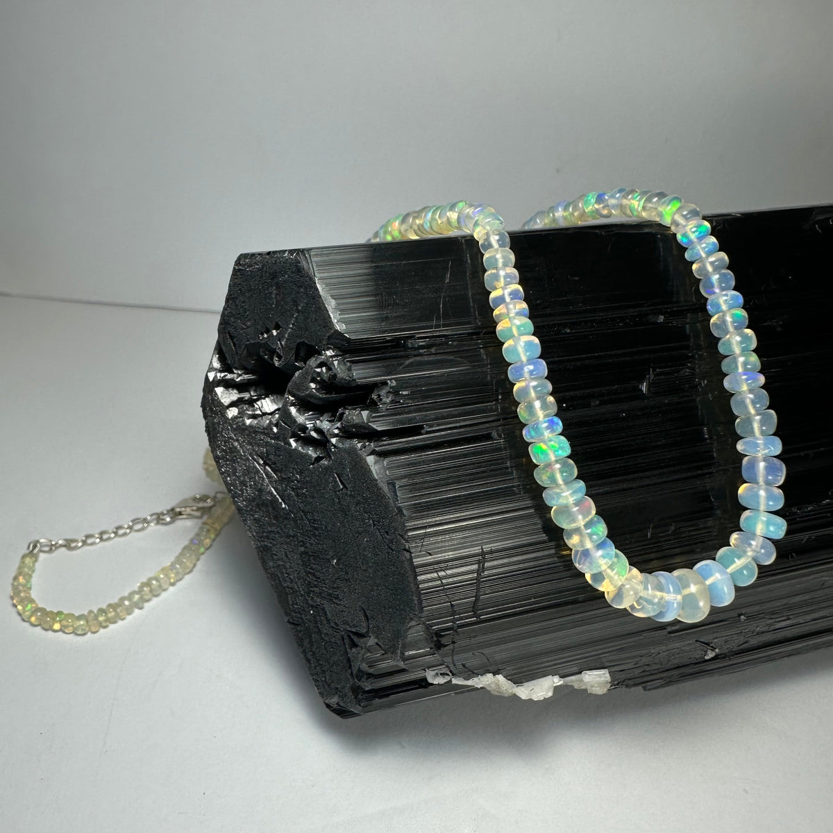 Graduated translucent opal beads with iridescent blue and green flashes strung into a necklace draped over a rough, shiny black tourmaline crystal cluster on a gray surface.
