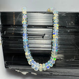 Graduated translucent opal beads in a necklace displaying iridescent flashes of green, yellow, and blue, arranged on a textured black surface.