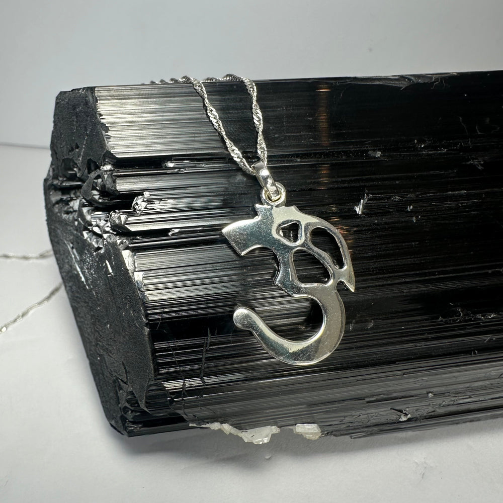 Polished silver 'Ohm' pendant with a stylized design hanging on a twisted silver chain, displayed on a black rough-textured crystal with striations.