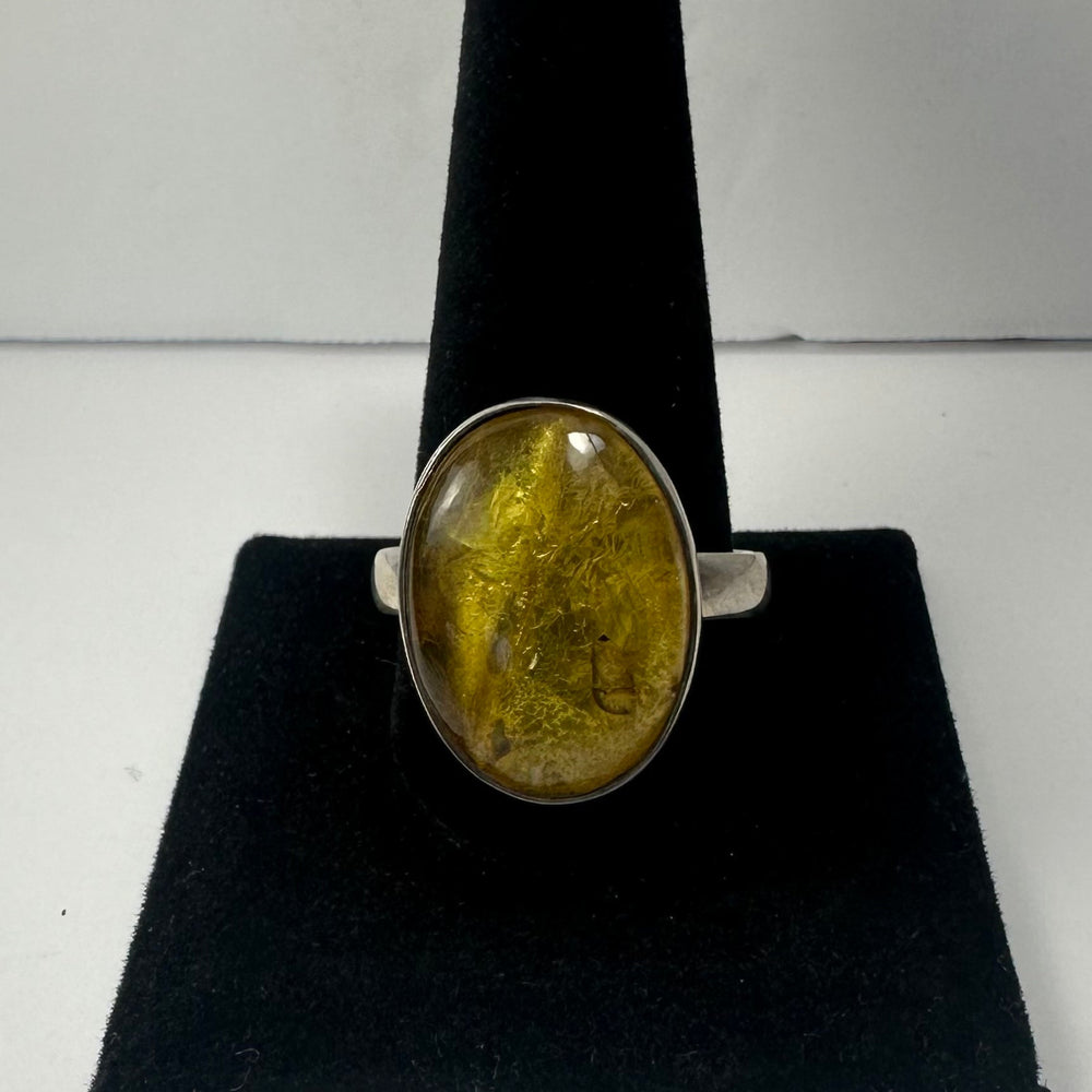 Oval amber stone ring with golden and brown inclusions set on a simple silver band, displayed on a black velvet ring holder against a plain background.