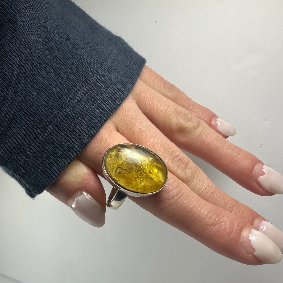 Hand wearing a silver ring with a large oval amber stone showing natural inclusions, against a plain light background and dark sleeve.