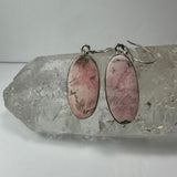 Oval pair of pink rhodonite stone earrings with black inclusions set in silver, displayed on a clear quartz crystal background.