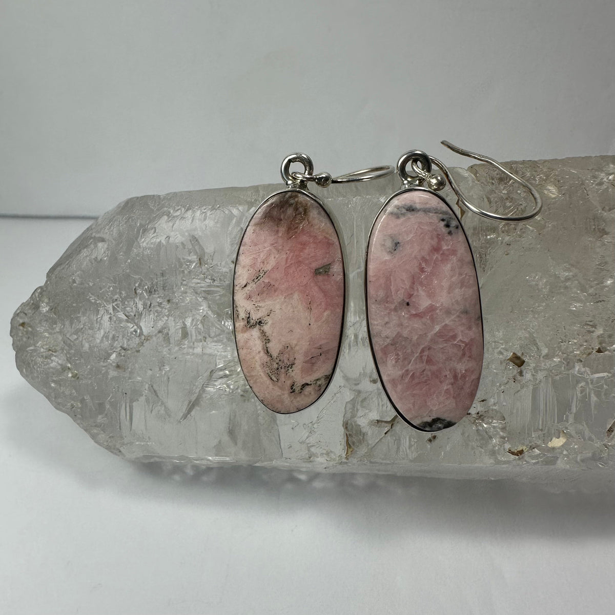 Oval pair of pink rhodonite stone earrings with black inclusions set in silver, displayed on a clear quartz crystal background.