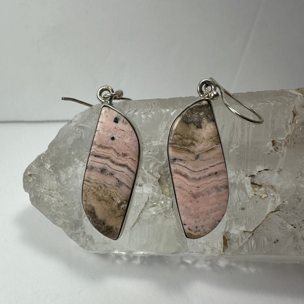 Pair of pink and brown striped rhodonite earrings set in silver, displayed resting on a rough clear quartz crystal against a plain background.