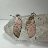 Pair of pink and brown striped rhodonite earrings set in silver, displayed resting on a rough clear quartz crystal against a plain background.