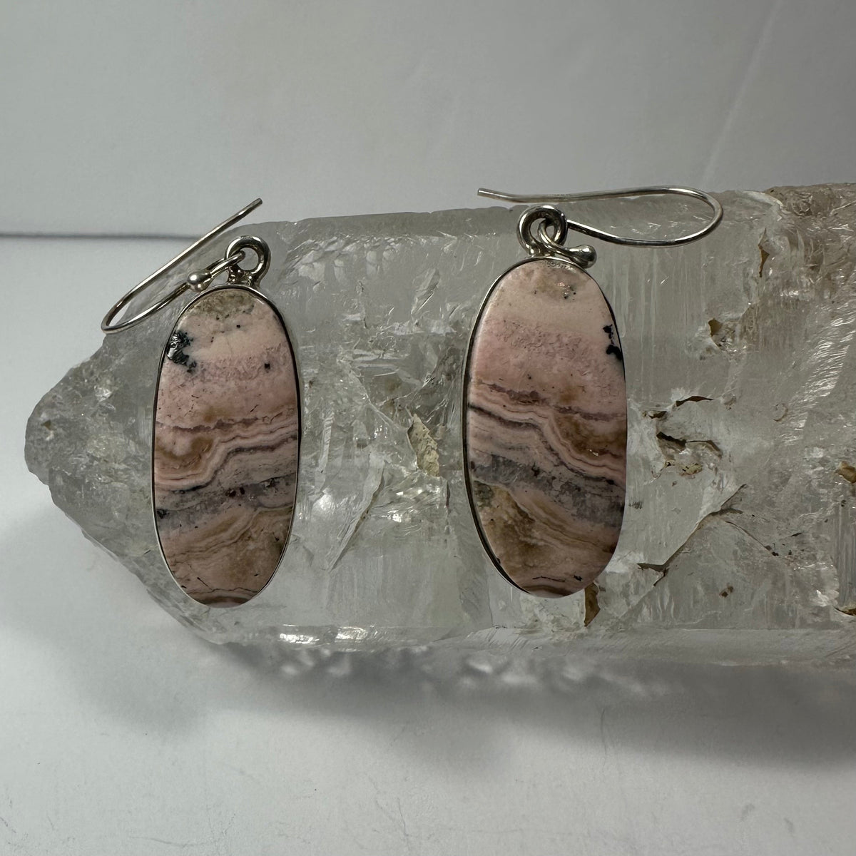Oval rhodonite stone dangle earrings in silver settings resting on a clear textured crystal cluster, showing pink and brown banded patterns.