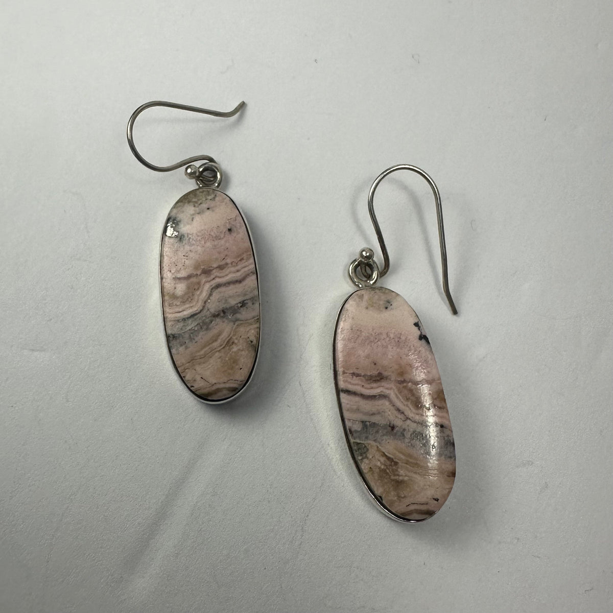 Pair of oval pink rhodonite stone dangle earrings with natural black and beige banding, set in silver metal with hook attachments on a white background.