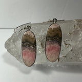 Oval-shaped rhodonite earrings with pink, gray, and beige bands set in silver, displayed resting on a clear quartz crystal cluster against a plain background.