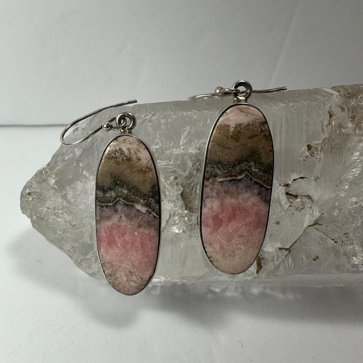 Oval-shaped rhodonite earrings with pink, gray, and beige bands set in silver, displayed resting on a clear quartz crystal cluster against a plain background.