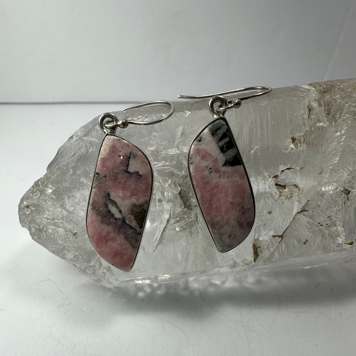 Pair of asymmetrical teardrop-shaped rhodonite stone earrings with pink, black, and gray marbled patterns set in silver, hanging on a clear quartz crystal.