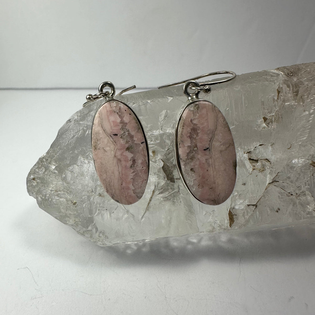 Oval-shaped Rhodonite earrings with silver hooks, displaying soft pink stones with black veining, resting on a clear quartz crystal cluster.