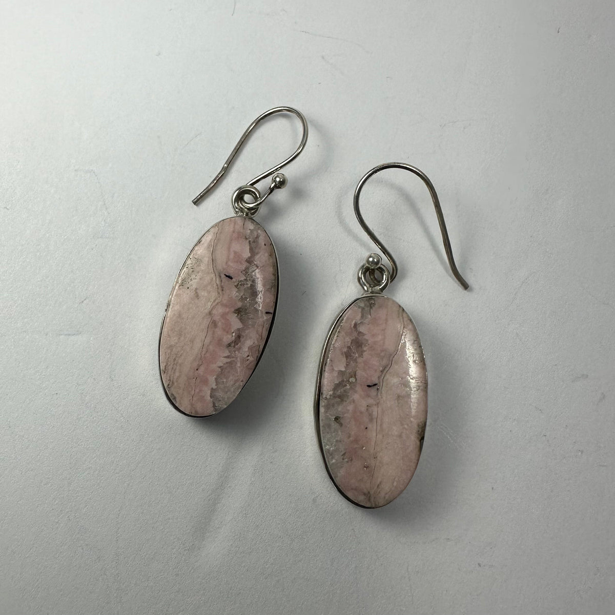 Pair of oval Rhodonite earrings with pink and black marbled patterns set in silver hooks against a plain white background.