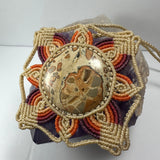 Hand woven beige, orange, purple, and red macrame flower necklace with a round jasper cabochon at center, placed on a rough purple crystal background.