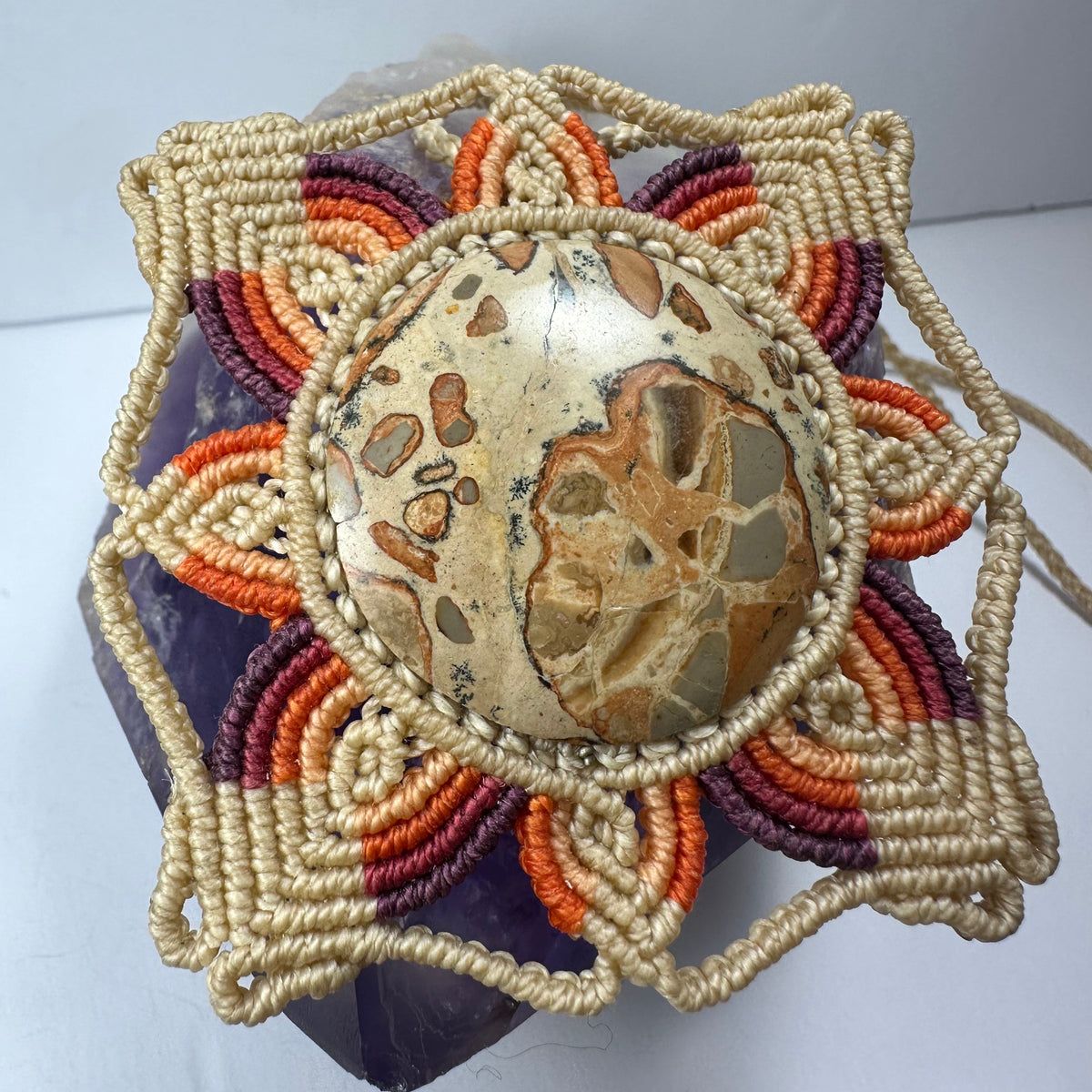 Handwoven flower-shaped necklace with concentric petals in cream, purple, and orange surrounding a round jasper stone with tan and brown patterns.