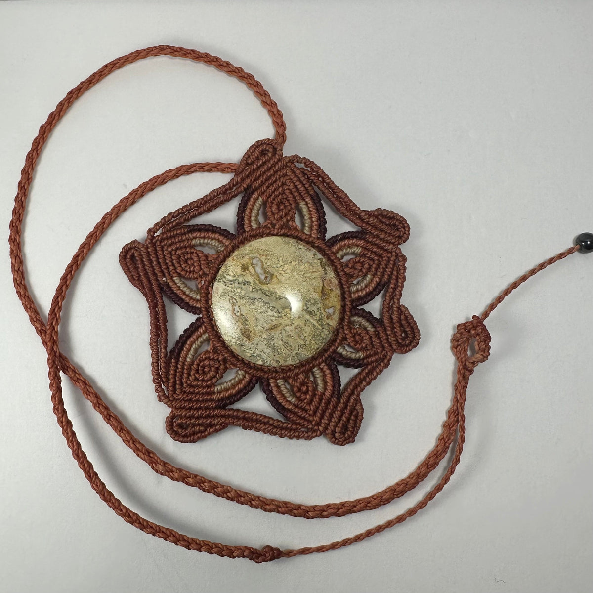 Handwoven necklace featuring a round jasper cabochon centerpiece framed by intricate brown cord in a flower-shaped pattern, with braided cord extending outward.
