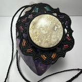 Round jasper cabochon with cream and beige patterns set in a black hand-woven macrame flower-shaped necklace with subtle teal, purple, and brown accents, resting on a purple crystal base.