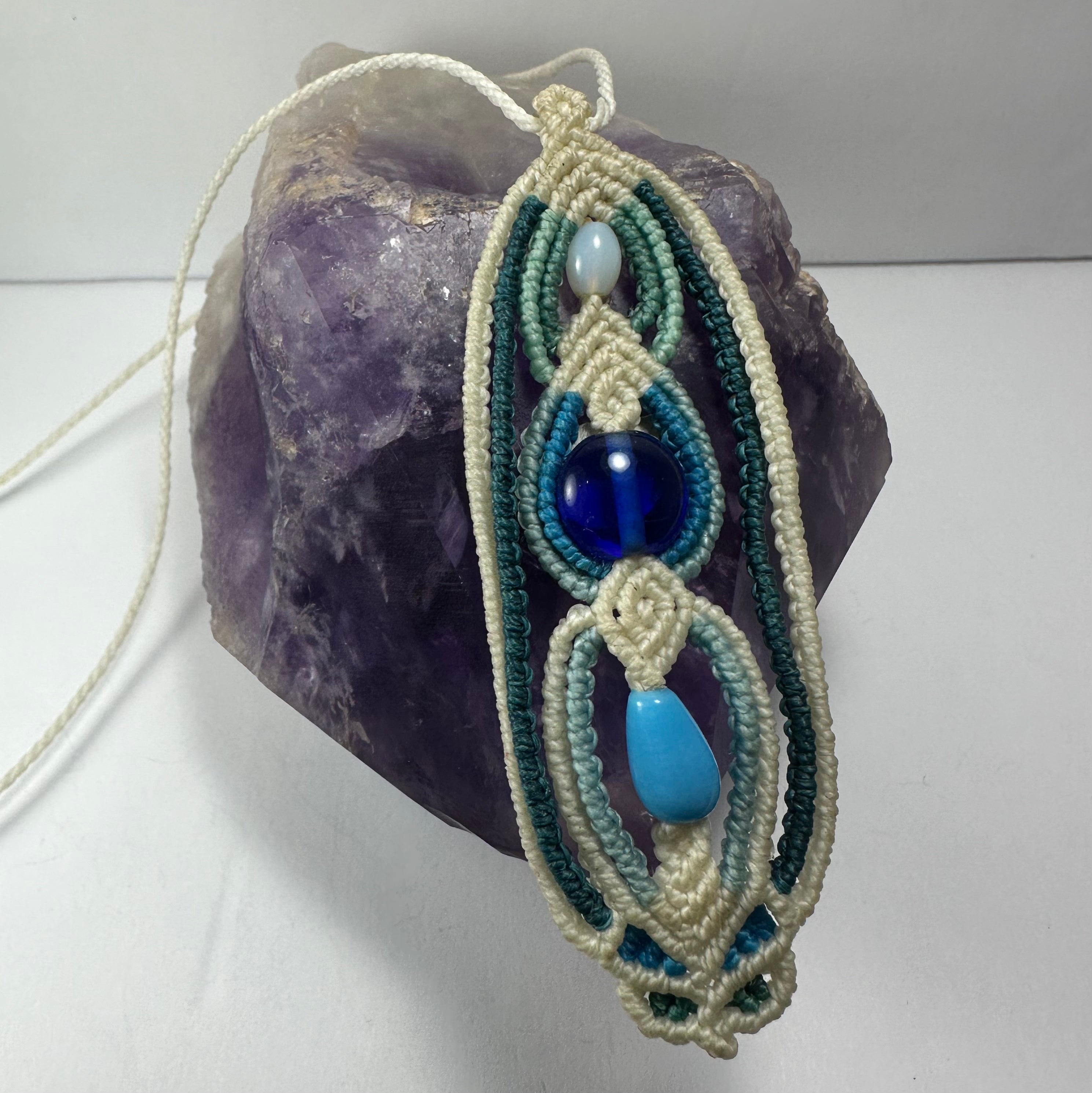 Hand-woven macrame necklace featuring glass beads and a 3.5 inch pendant displayed on fabric.