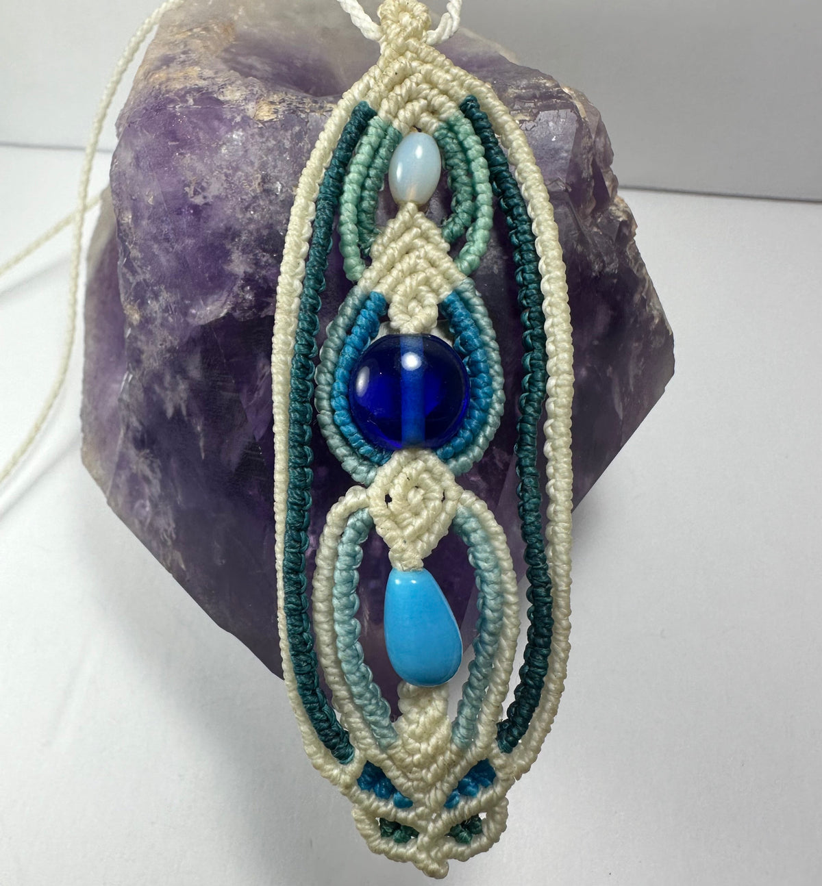 Hand-woven macrame necklace featuring cream, teal, and blue cords with a large central dark blue glass bead and smaller white and light blue beads, resting on a purple amethyst stone.