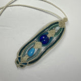 Hand-woven macrame necklace pendant in beige cord with a central dark blue glass bead, a teardrop-shaped light blue bead, and additional small round beads, on white background.