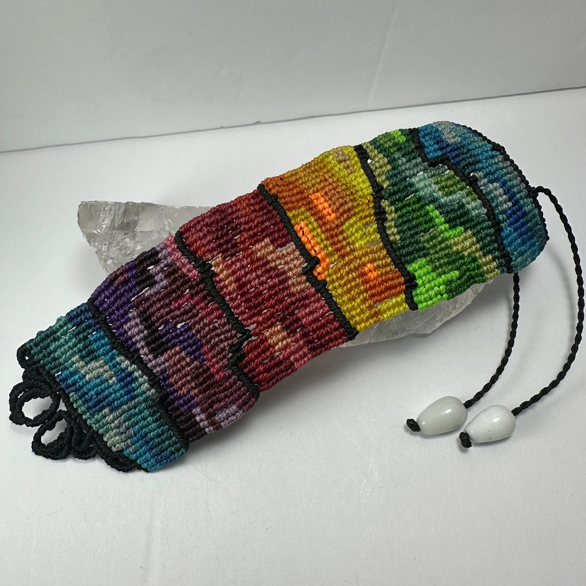 Handwoven bracelet with horizontal stripes transitioning through blue, purple, red, orange, yellow, green, and teal, separated by thin black lines with a black braided closure and white beads.