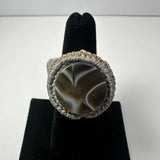 Round ring featuring a polished brown and white patterned stone encased in a handmade light beige woven band, displayed on a black velvet ring holder.