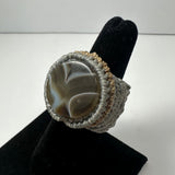 Round polished stone with white and brown bands woven into a wide taupe and beige macrame ring band, displayed on a black velvet ring stand.