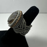 Handwoven ring featuring a polished round brown stone set in tightly braided beige and gray fibers, displayed on a black velvet ring holder against a plain background.