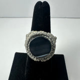 Handmade woven ring with a large smooth black circular stone in the center, surrounded by tightly braided light gray fiber, displayed on a black velvet ring stand.
