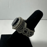 Handmade ring with a polished black stone set in intricately woven beige cord bands, displayed on a black velvet ring holder against a gray background.