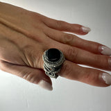Hand wearing a handmade woven ring with a round black stone centerpiece and intricate gray cord weaving on a white background.