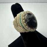Handwoven cream-colored ring with teal accents featuring a round black and gray marbled stone centerpiece, displayed on a black ring stand.